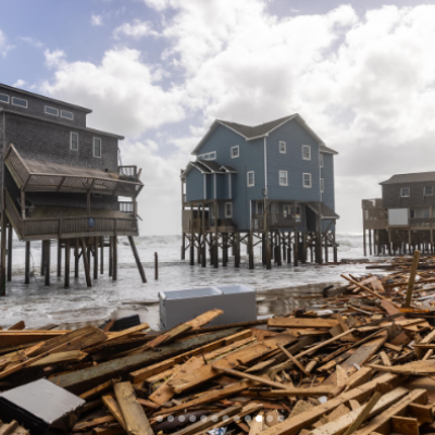 Stilt houses by the sea surrounded by debris and scattered wood under a cloudy sky.