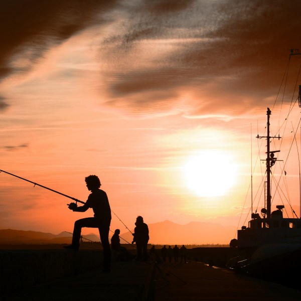 Silhouette of people fishing on a dock at sunset with a boat nearby.