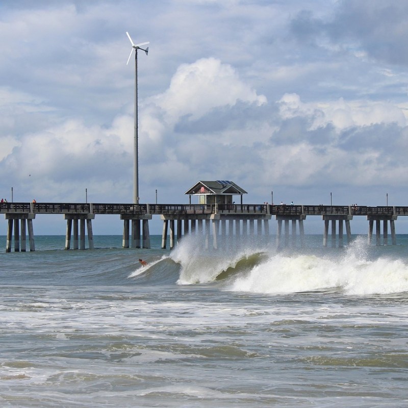 Surfer riding a wave near a long pier with wind turbines and cloudy sky in the background.