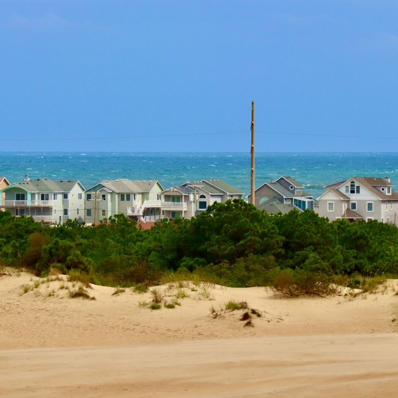 Row of beach houses behind sand dunes and vegetation, with ocean in the background.