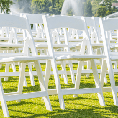 Rows of white folding chairs on green grass with sprinklers in the background.