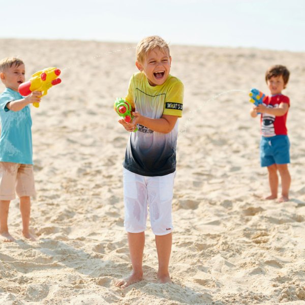 Three boys playing with water guns on a sandy beach.