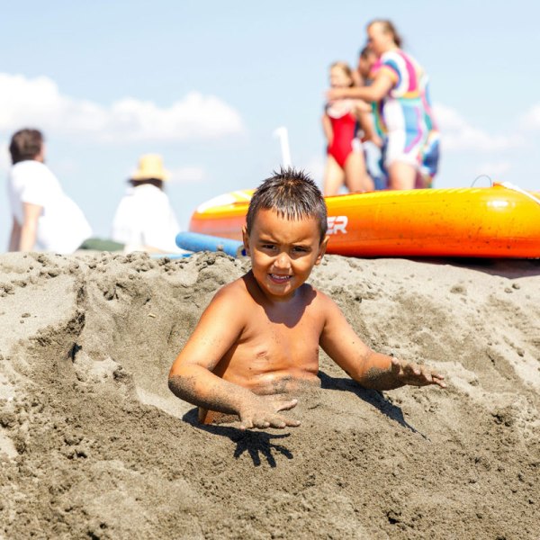 Child playing in sand with people and an inflatable in background.