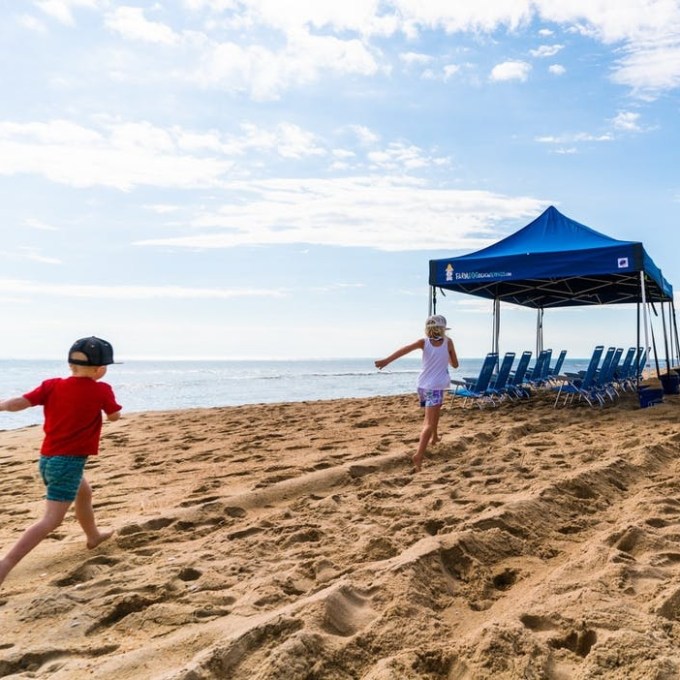 Two children run on a sandy beach near a blue canopy with chairs.