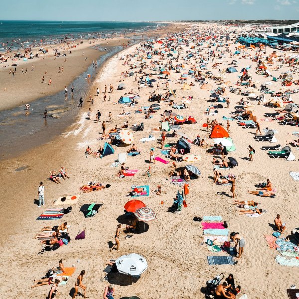 Crowded beach with sunbathers, umbrellas, and beach towels stretching along the shoreline.