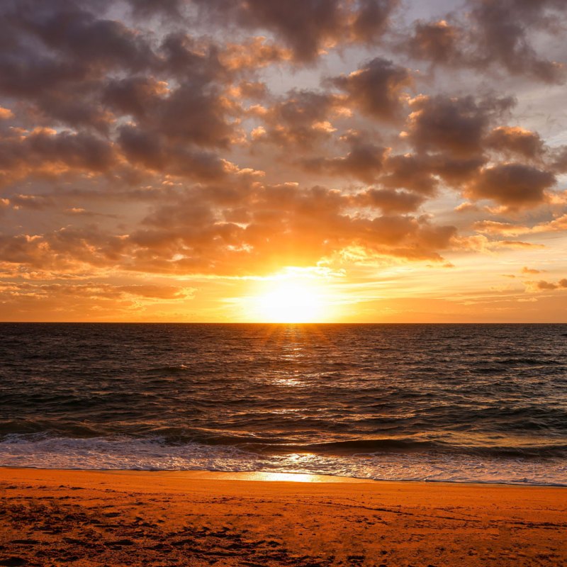 Sunset over ocean with cloudy sky and sandy beach in foreground.