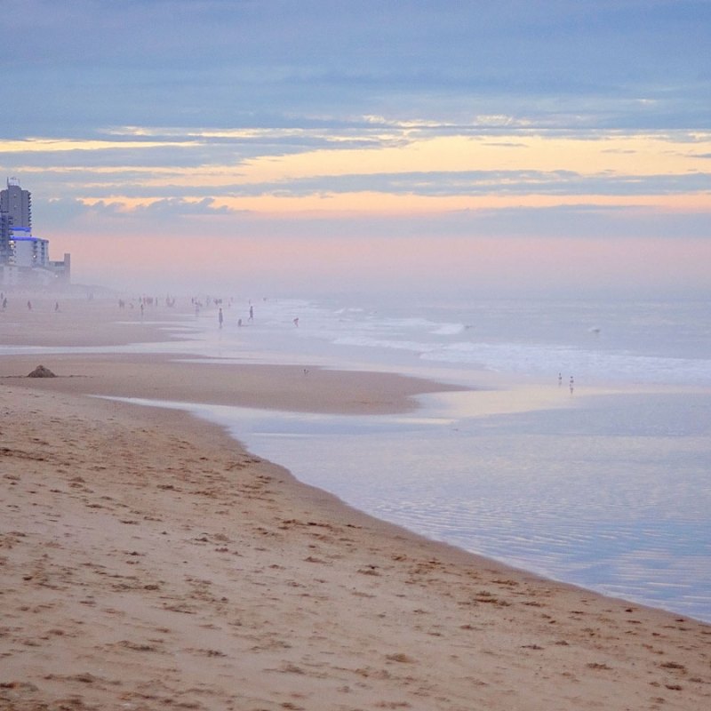 Beach at sunset with distant buildings, calm waves, and a pastel sky.