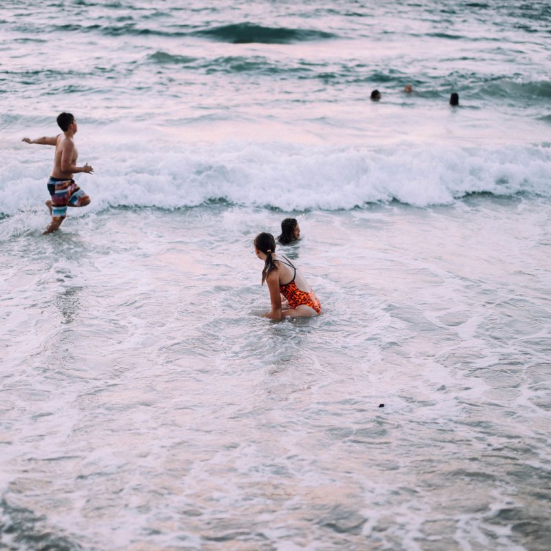 Kids playing in ocean waves at sunset with others swimming in background.